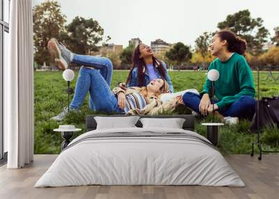 Three happy young women having fun sitting on green grass at park. Multi-ethnic group of female students laughing while relaxing at college campus Wall mural