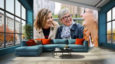 Three mature retired women laughing while talking sitting outside. Small group of mature females having fun enjoying a conversation bonding at city street. Elderly friendship concept. Wall mural