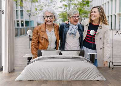 Three mature retired women laughing while walking together outside. Small group of 60s females having fun enjoying a conversation bonding at city street. Elderly friendship concept. Wall mural