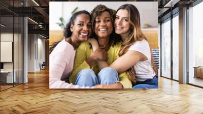 Three united beautiful smiling women sitting together on couch - Portrait of young multiracial girl friends hugging each other and relaxing at home - Friendship, youth and millennial people concept Wall mural