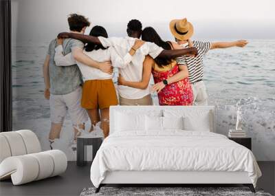 View from back of group of young diverse people friends hugging each other and enjoying sunset at beach in summer holidays Wall mural