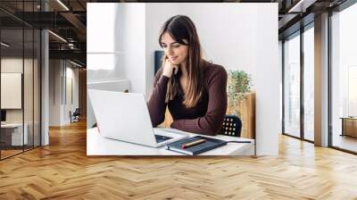 Young female student learning online in living room. Millennial woman sitting on table listening online training class while studying remotely from home. E-learning and education concept. Wall mural