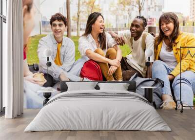 Young group of multiracial students laughing and having fun together sitting on the grass in the university campus. Teenage classmates social gathering relaxing outdoors. Wall mural