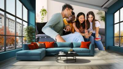 Young group of people using mobile phone device sitting together on sofa at home. Inclusion and diversity concept with young indian man in wheelchair having fun with friends watching content on phone. Wall mural