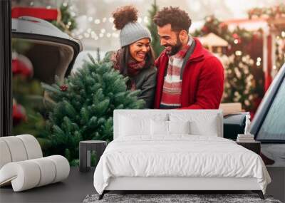 A young couple is loading a Christmas tree into the trunk of their car, smiling and laughing together as they prepare to move home for the holiday season. Wall mural