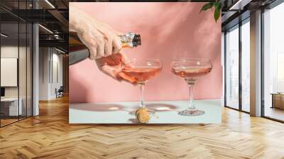 Female hand pouring champagne or wine into glasses. Gentle pink background bright sunlight. Copy space minimalism. Wall mural