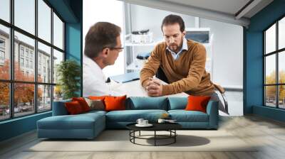 Adult doctor speaking with bearded man in his office while recording necessary data on paper Wall mural