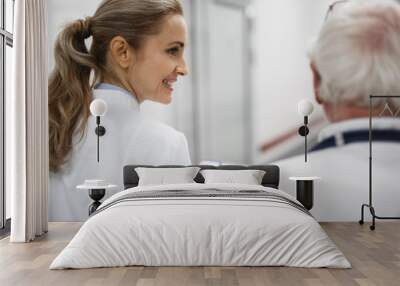 Back view portrait of beautiful young lady in white lab coat holding clipboard and chatting with old therapist. They heading to patient room Wall mural