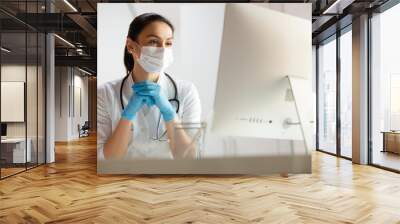 Beautiful doctor in medical mask doing tasks at laptop Wall mural