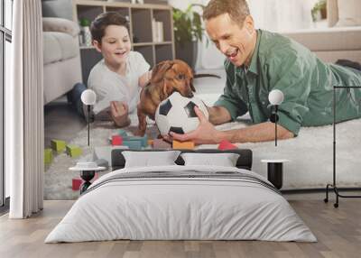 Cheerful dog is biting ball in male hands playfully. Father and son are looking at pet and smiling Wall mural