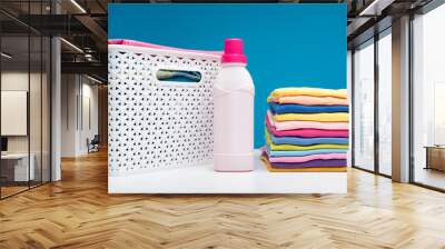 Close up of colorful garments stacked in pile. Bottle of softener and wicker clothesbasket are standing aside Wall mural