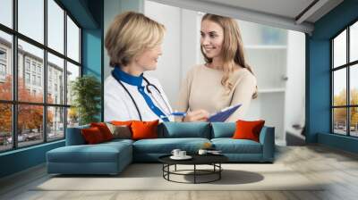 Concept of professional consultation in healthcare system. Waist up portrait of smiling female doctor talking with patient while writing in her notes holder Wall mural