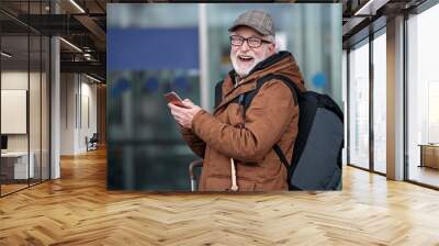 Fanny moments. Laughing old gray-haired man with beard is standing outdoors with backpack and mobile phone. He is looking at camera with wide smile Wall mural