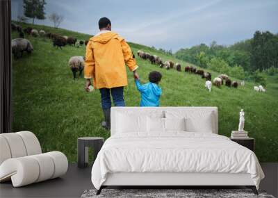 Father and son wearing rubber boats and raincoats going hand by hand at the green hill Wall mural