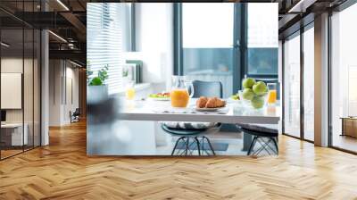 Fresh orange juice in jug, croissants and apples on table in kitchen. Plates with salad on background Wall mural