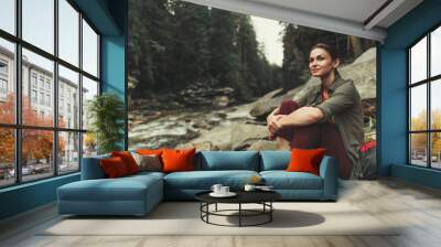 Friendly young woman sitting on the rocks of the mountain river while enjoying beautiful views Wall mural