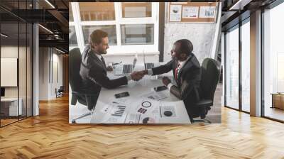 Great job. Top view of stylish young businessmen are sitting at workplace opposite and looking at each other with smile while shaking hands Wall mural