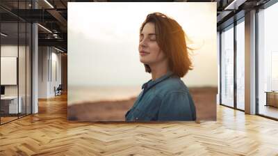 Great pleasure. Charming calm young woman is standing near sea with closed eyes and expressing delight. She is posing against wonderful sunset while enjoying last rays of the sun Wall mural