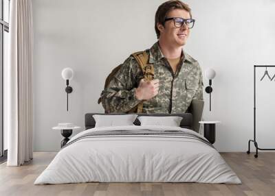 Happy male cadet holding laptop and smiling. He is wearing eyeglasses and having rucksack on shoulder. Isolated on background Wall mural