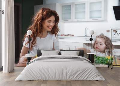 Hungry little girl is eating tasty food with appetite while sitting at table in kitchen. Her mom is laughing  Wall mural