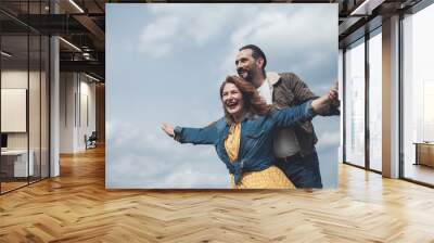 I am free. Portrait of excited woman is stretching arms sideways while man is embracing her from behind. They are enjoying wind and sky together  Wall mural