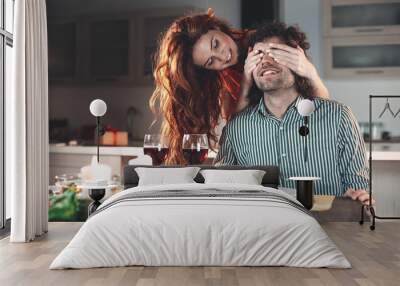 I have surprise for you. Positive girl is closing eyes of guy secretly and smiling. Loving couple is having romantic dinner in kitchen  Wall mural