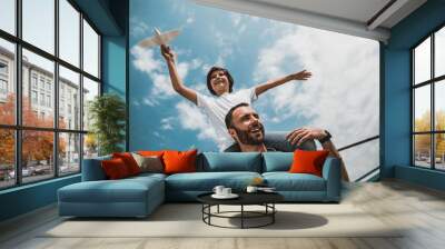 Low angle portrait of laughing bearded man and positive kid having fun with plastic toy under blue sky with white fluffy clouds Wall mural