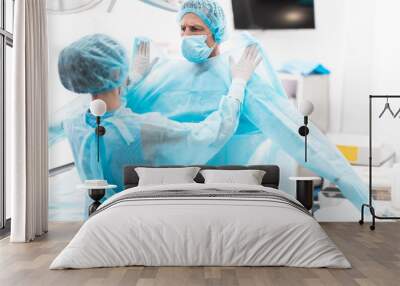 Medical workers. Portrait of nurse in sterile gloves putting on blue surgical gown on doctor Wall mural