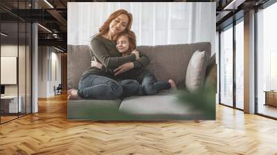 My dearest. Full length portrait of mother and her child hugging each other and sitting on couch at home. They are smiling, calm and happy Wall mural