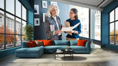 My report. Confident serious gray-haired businesswoman is standing with her young professional employee against window. Positive girl is showing folder with documents to her qualified chief Wall mural