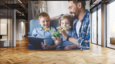 Outgoing dad having fun with satisfied children. They looking at toy. Glad boy typing in gadget Wall mural