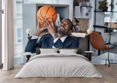 Playful mood. Cheerful young african manager in suit is sitting in office and holding orange basketball with smile. He is looking up while being ready to throwing it Wall mural