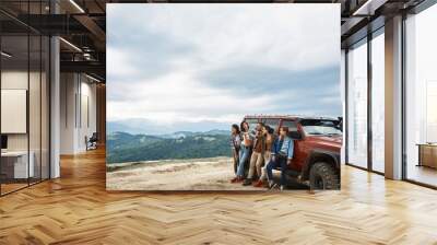 Pleasant happy young friends standing near their car while enjoying view from the mountain hill Wall mural