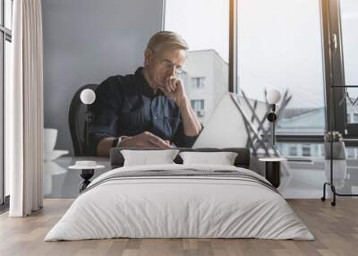 Portrait of concentrated old businessman looking at laptop while sitting at table in office. Abstracted worker during occupation concept Wall mural