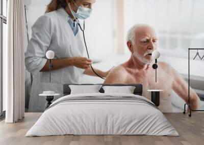 Portrait of doctor in protective mask checking patient breath with stethoscope. Shirtless bearded gentleman sitting on hospital bed Wall mural