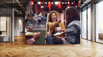 Portrait of smiling young lady in hat sitting on folding chair and letting boyfriend taste french fry. Young people resting during outdoor concert Wall mural