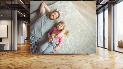 Relaxing at home together. Top view of joyful dad is lying of soft carpet and embracing his little daughter. They are looking at camera and laughing. Copy space Wall mural