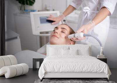 Side view of happy young woman getting cavitation rejuvenating skin treatment at spa. She is lying on massage table and smiling. Beautician is touching monitor screen while holding tool near female Wall mural