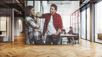 Side view profile of optimistic girl is drinking espresso while leaning on glass wall. She is looking at man who is standing near and flirting with her. Professional team is laboring in background Wall mural