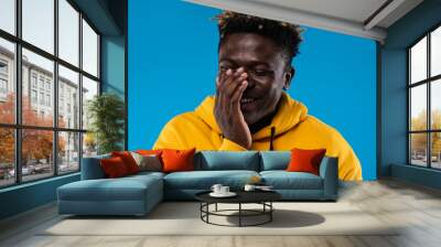 Stop me. Close up portrait of cheerful guy with closed eyes covering face with hand. He is wearing yellow hoodie and standing on blue background Wall mural