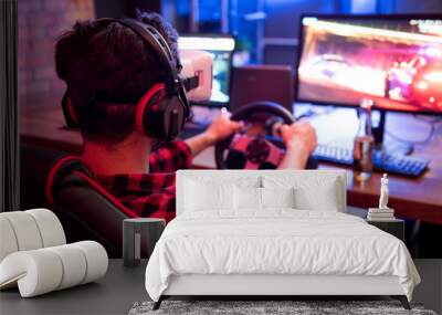 Three-dimensional image. Back view of young man is enjoying car racing video game while sitting in chair wearing virtual reality goggles and holding steering wheel. Table with monitor in background Wall mural