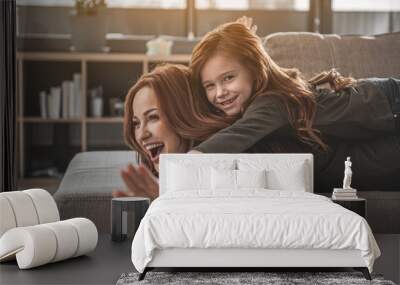 Up. Waist up portrait of laughing red-haired woman and smiling daughter playing together. Small girl is lying on her mother and holding hands as if being bird Wall mural