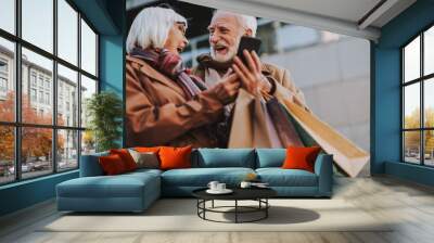 Waist up portrait of cheerful old lady holding cellphone while husband with shopping bags looking at her and laughing Wall mural