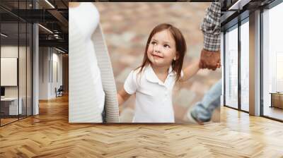 Waist up portrait of little kid looking at mom with joy. She is walking with mother and father holding hands and feeling delighted Wall mural