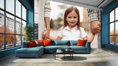 Waist up portrait of small smiling girl standing and holding mother and father hands. She is happy to spend time with dearest together Wall mural