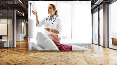 Waist up portrait of young woman in white lab coat checking intravenous drip in hospital room. Blurred old man lying in bed Wall mural