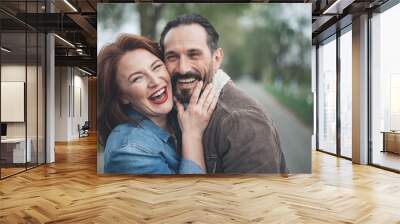 We are happy together. Joyful husband and wife are bonding to each other with fondness. They are looking at camera and laughing while standing on path outdoor  Wall mural