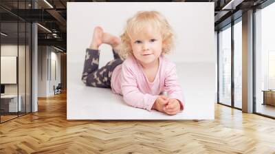 curly blonde baby girl in comfortable home casual clothes is lying on the floor in a bright white room smiling and looking at the camera Wall mural