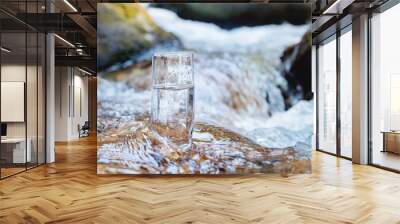 A glass glass with pure mountain drinking water stands on a rock in the course of a mountain river against the backdrop of seething cascades and waterfalls of a mountain river. The concept of pure Wall mural