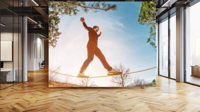 a man, aged with a beard and wearing sunglasses, balances on a slackline in the open air between two Wall mural